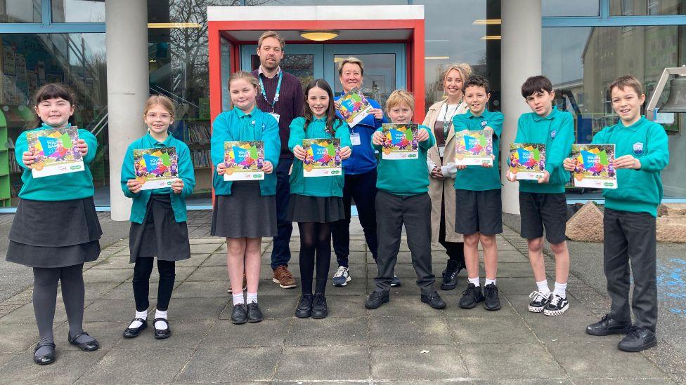 Three adults and a group of schoolchildren with cyan tops hold out a book with a colourful cover