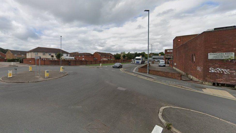 Roundabout with three roads leading to it - houses are along the far side and a brick building stretches along the right side.