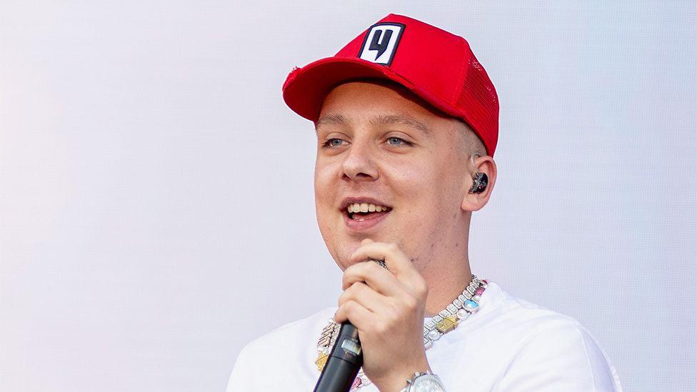 Close up of Aitch's face wearing a red cap holding a microphone 