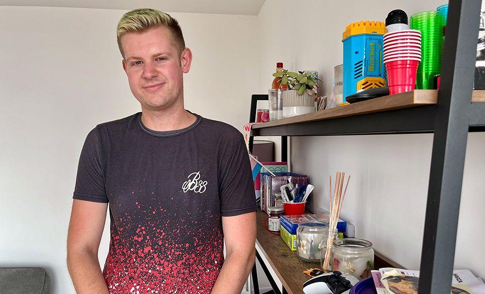 Guy Benson stands in his shared ownership flat at Landmark Pinnacle, he's wearing a black and red T shirt and smiling with blonde hair, next to a wall and a set of shelves with various homely possessions on them.