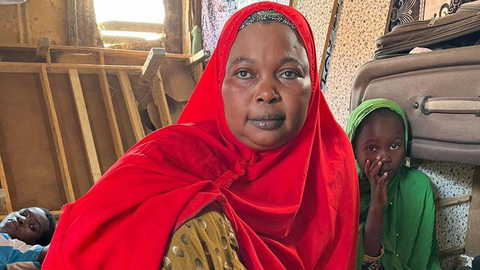 Hawa Mohamed Zubery wearing a red headscarf and pictured sitting on the floor in her dwelling in Eyl with two of her children looking on from the either side