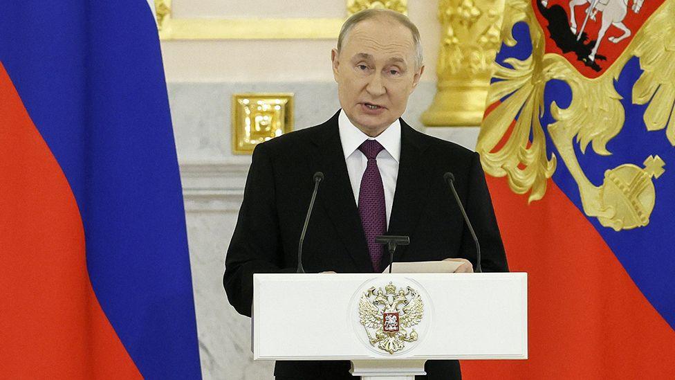 Vladimir Putin stands at a lecturn wearing a suit and purple tie with the Russian flag behind him to the left, in Moscow on 5 November (file photo)