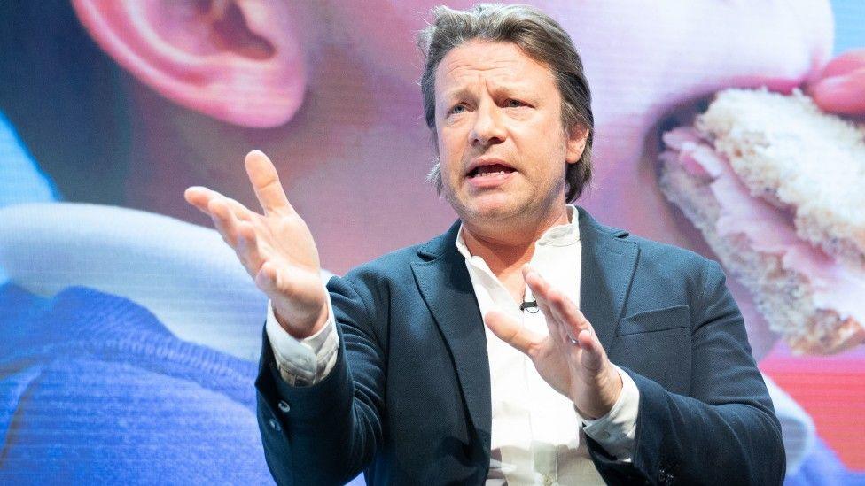 Chef Jamie Oliver at a public speaking event, gesticulating with his hands, in front of a screen showing a child eating a sandwich