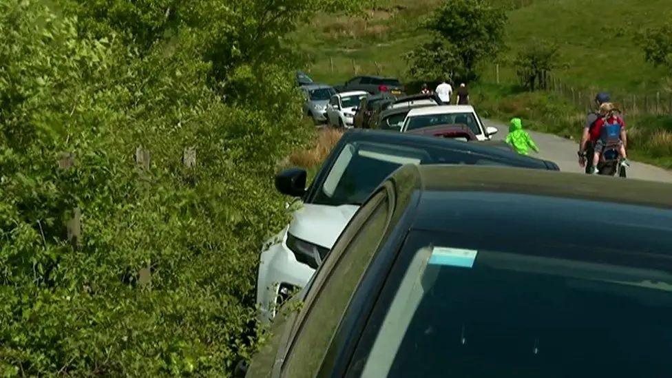 Cars parked on either side of a verge on a narrow road.