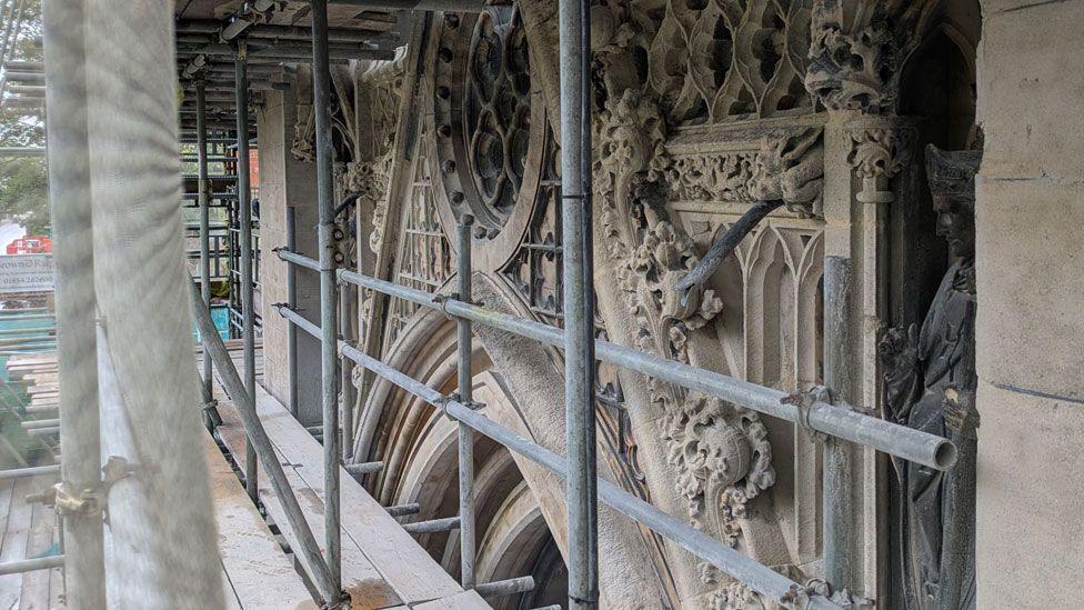 A side on close-up view of some of the elaborate Victorian gothic style carving on the Church of Our Lady and the English Martyrs. It shows the top of an arched window, above it a round window. In font of it are grey scaffolding poles and a wooden walkway.