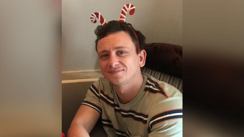 A man with short brown hair is smiling and wearing a candy cane headband and a beige top with black and white stripes. He is sitting indoors on a chair.