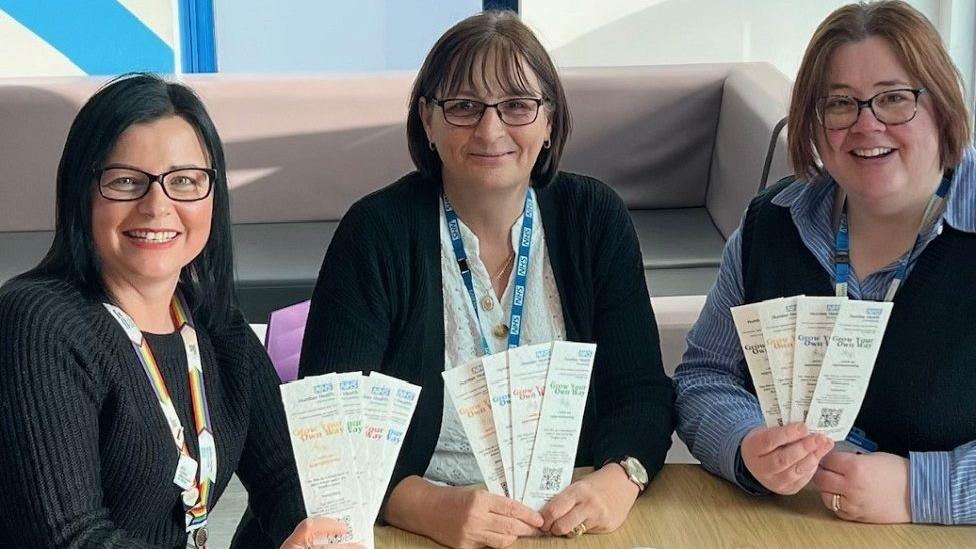 Three female staff members, all with glasses and short dark hair with lanyards around their neck, holding leaflets promoting nursing apprenticeships.