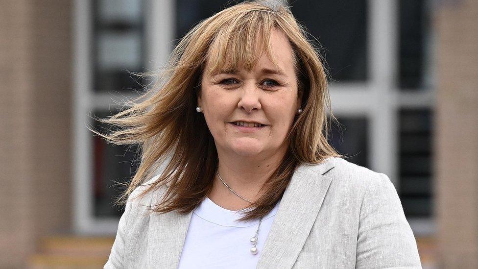 Michelle McIlveen pictured in a white shirt with grey suit jacket, wearing a silver necklace, she has shoulder length brown hair and looks windswept