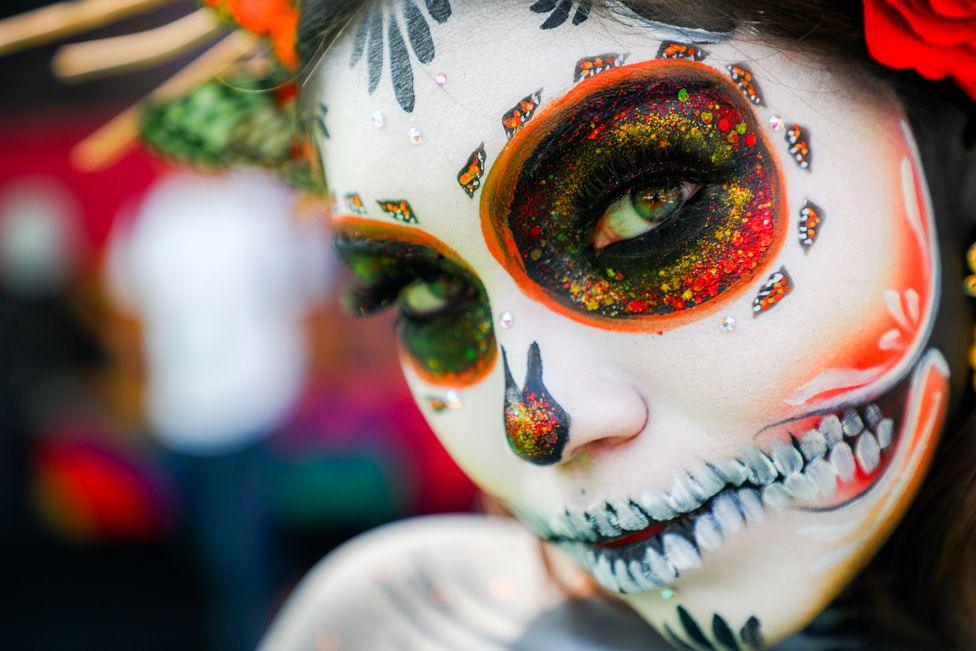 A close up shot of a woman wearing day of the dead make up of a La Catrina, a skull grin in vivid hues of black, white and orange