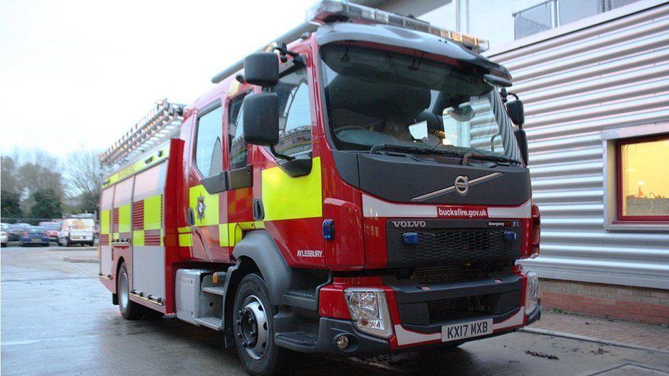 A Buckinghamshire Fire & Rescue Service fire engine. It is sideways on, with the majority of its empty cab on the right side and the body of the engine receding into the left side. It is mostly painted bright red with yellow and on its cab, beneath the window, can be seen a sign saying bucksfire.gov.uk