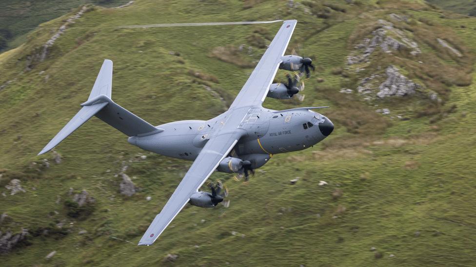 The A400M in the air, flying over a green, rocky, hilly region.