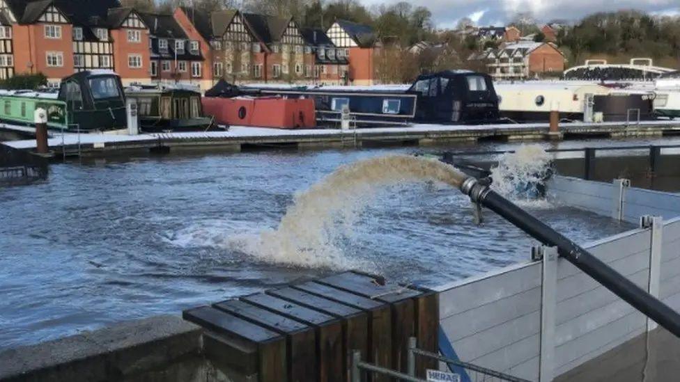 A pump ejects water out from over a flood barrier in Northwich, with a jetty, canal boats and homes seen in the distance. 