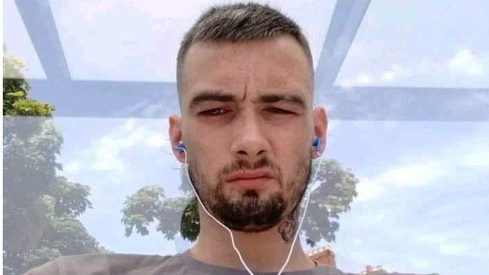 A close-up photo of the late Ryan MacNab.  He has short hair and a beard.  He is wearing a small set of earphones and a grey t-shirt.  There is a view of a cloudy blue sky and trees out of the window behind him.