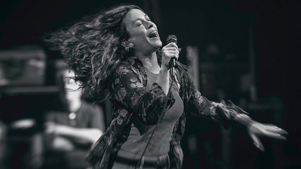 Alanis Morissette in a black and white photo, singing on stage with her eyes closed and her long hair streaming behind her