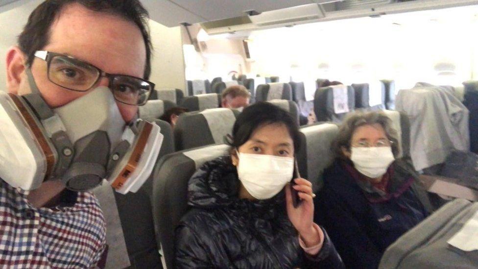 A man wearing a face mask looks into the camera in a selfie style, while two women wearing face masks also look at the camera, sitting on plane seats