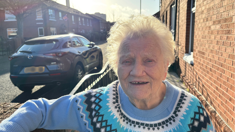 Margaret Totton. She has short white hair, wearing a blue fair isle jumper. Her arm is resting on a railing. She is standing on a residential street with houses in the background.