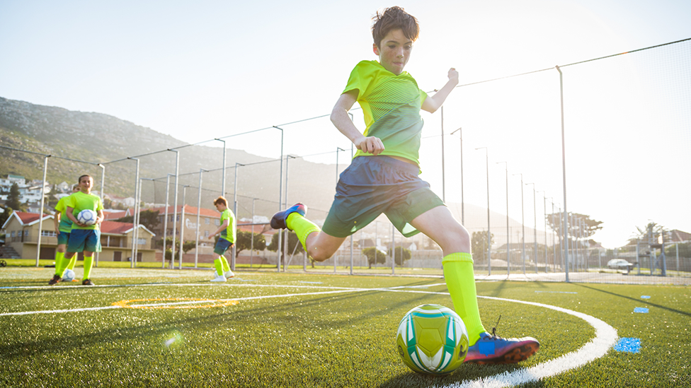 A young boy goes to take a kick on a football pitch with other children in the background. He's wearing a yellow and blue strip. The setting for the pitch looks like a valleys community.