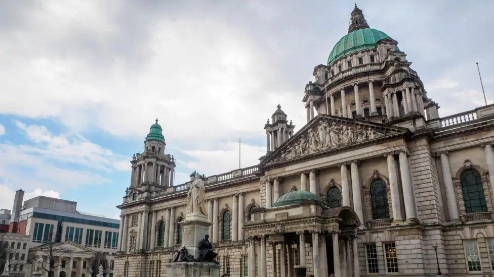A picture of Belfast City Hall. It is a grand building with faded white stone and three towers each of which have a turquoise metal roof.