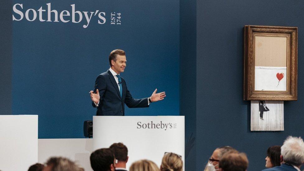 The Sotheby's auction at which the renamed Love Is In The Bin piece by Banksy was sold. There is an auctioneer standing behind a podium at the front, in front of a large blue wall. He is wearing a dark suit and blue tie with his arms outstretched as he collects bids. On the right the artwork is displayed. It is a spray painted girl reaching for a red heart-shaped balloon. The piece has been shredded and is half-hanging out of its gold frame.