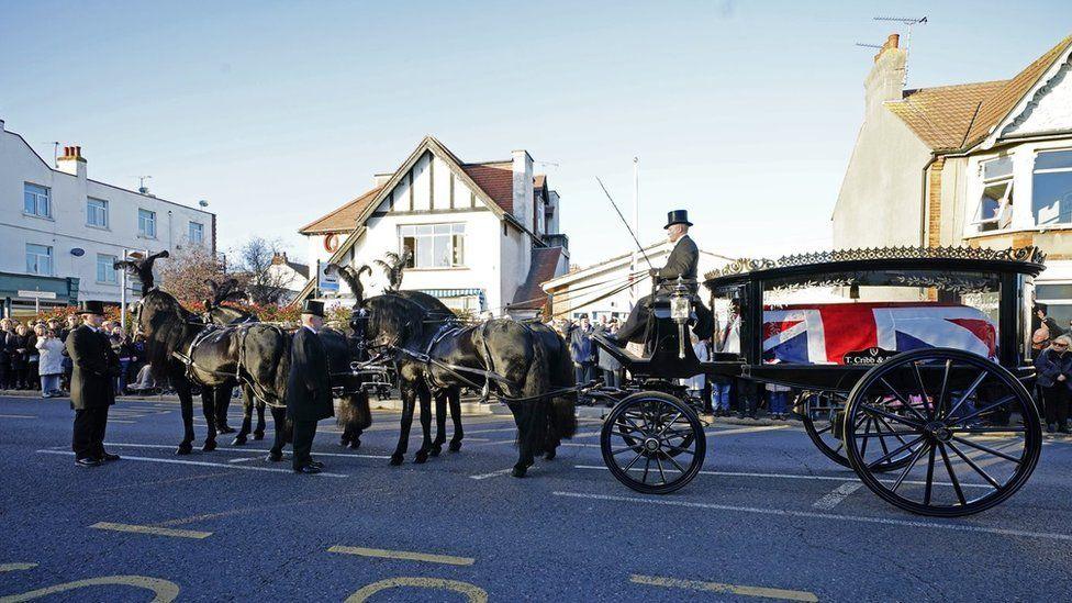 Four black horses pulling a carriage with a coffin inside draped in the union flag. Crowds are gathered in the background and watching the procession