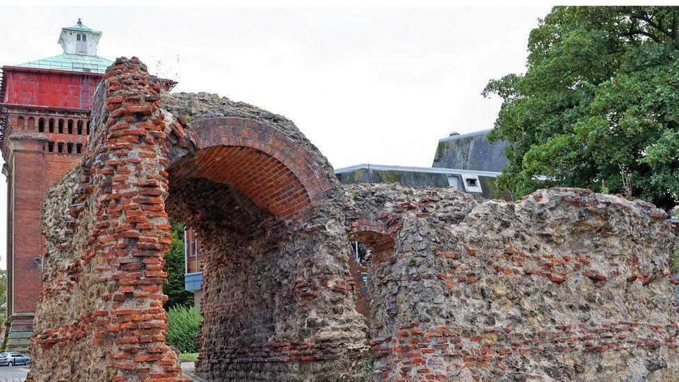 Balkerne Gate in Colchester. It is a red brick wall that has crumbled in parts. There is an archway in the wall.