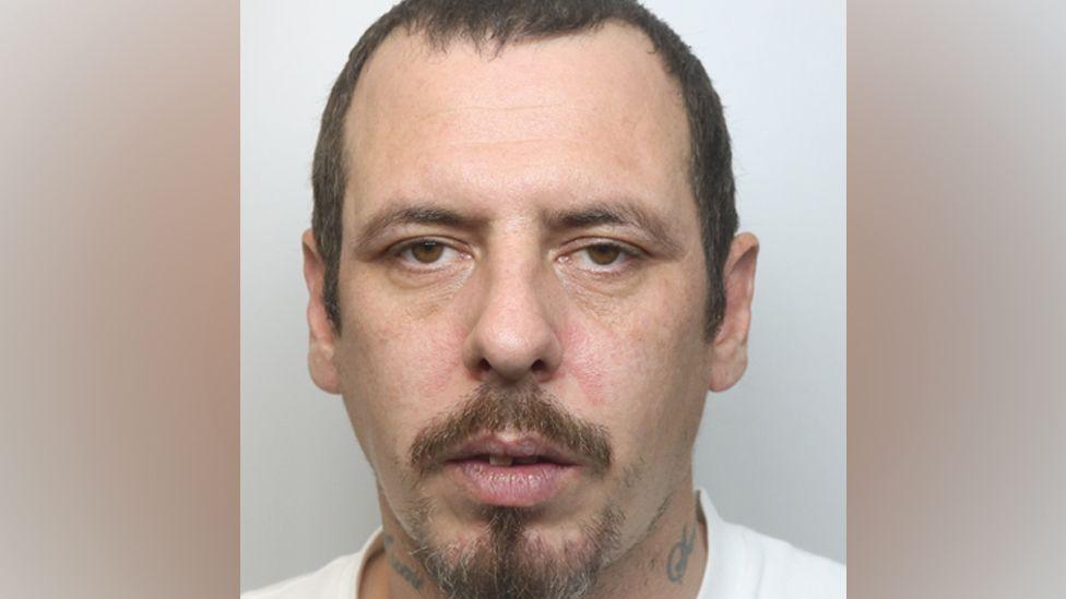 A police custody photo of Kieron Stuart Robert Long looking at the camera, with his mouth slightly open. He has short dark hair, a goatee beard and tattoos on his neck. He is wearing a white T-shirt.