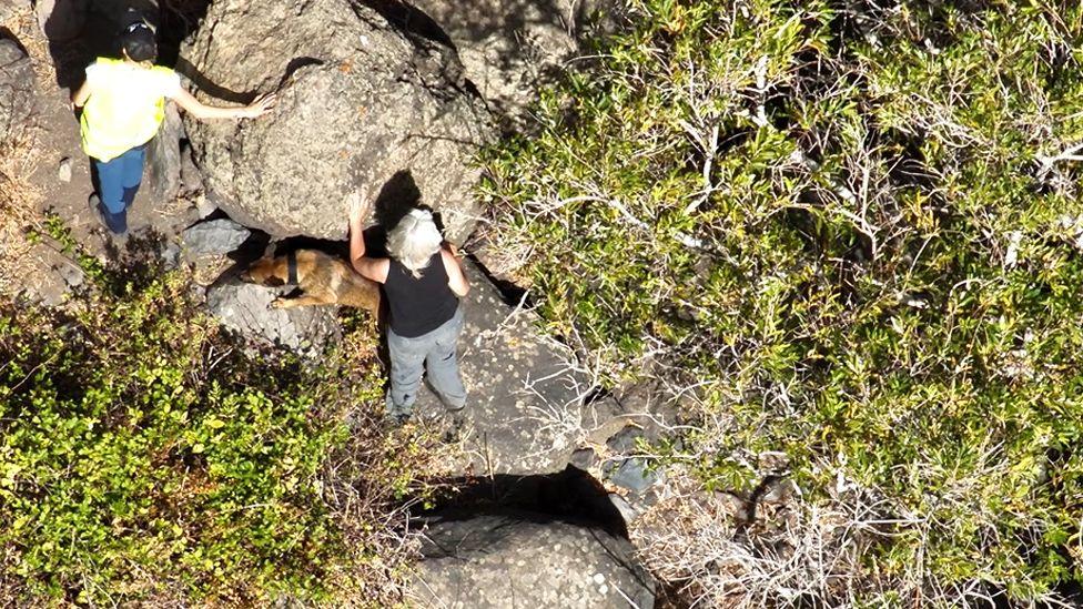 Aerial image of two women and a dog searching among rocks and scrub