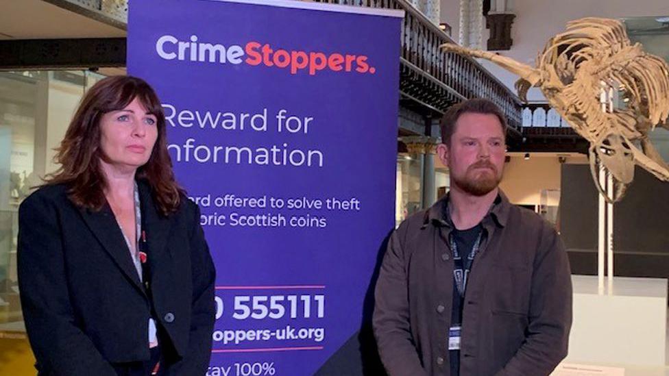 A woman with long hair and a dark jacked and a man with red hair and a beard stand either side of a poster for Crimestoppers offering a reward for information