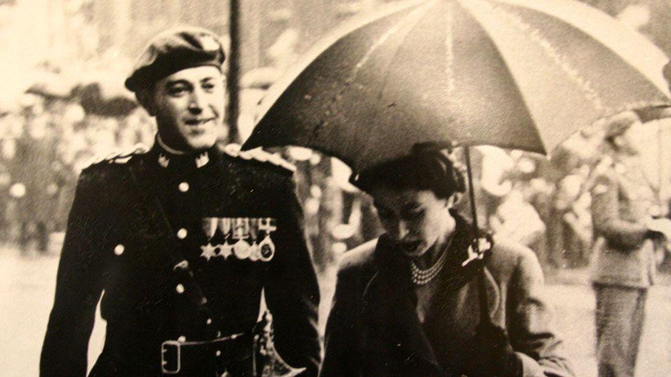 A black and white image showing John Beckett with Queen Elizabeth II in 1955. He is on the left wearing a military uniform with an array of medals on his left chest and a beret and is looking towards the queen on the right. She is holding an umbrella up, is looking down and is wearing a thick coat or jacket and a string of pearls. Behind them soldiers can just be seen lining up. 