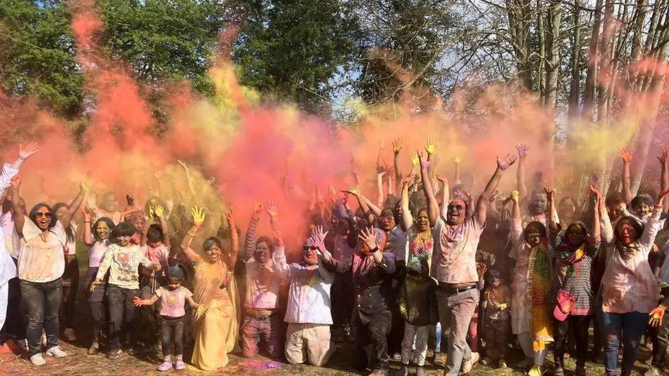 A group of people in a line men, women and children. They are throwing colourful powders into the air, with colours including pink, yellow and orange. 