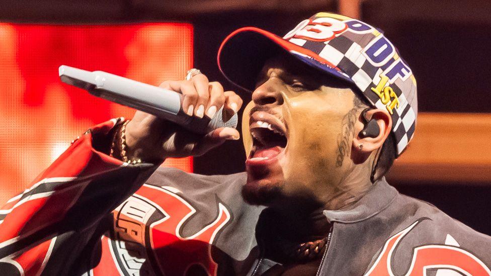 Chris Brown in a red, white and black patterned leather jacket and patterned baseball cap, singing into a microphone on stage at the Tycoon Music Festival at Little Caesars Arena on April 19, 2025 in Detroit, Michigan.