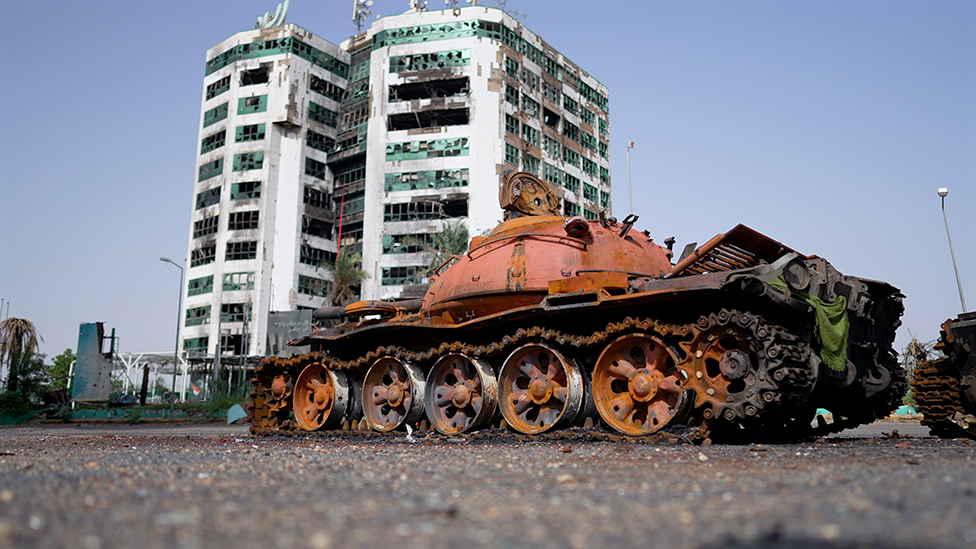 A rusted tank sits on the road in front of a ruined high-rise building.