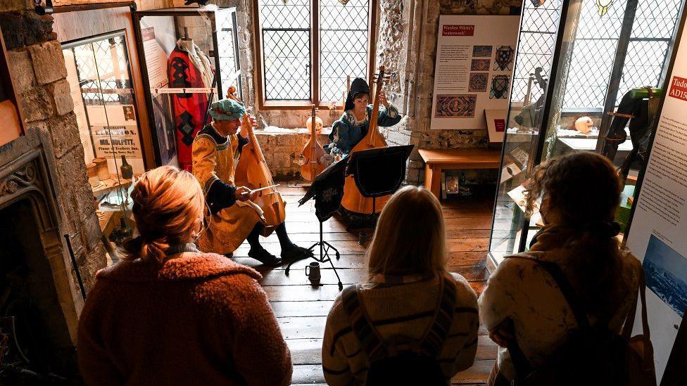 A group of three people watch two people sitting playing instruments while dressed in medieval clothing. They are all inside Westgate Museum.