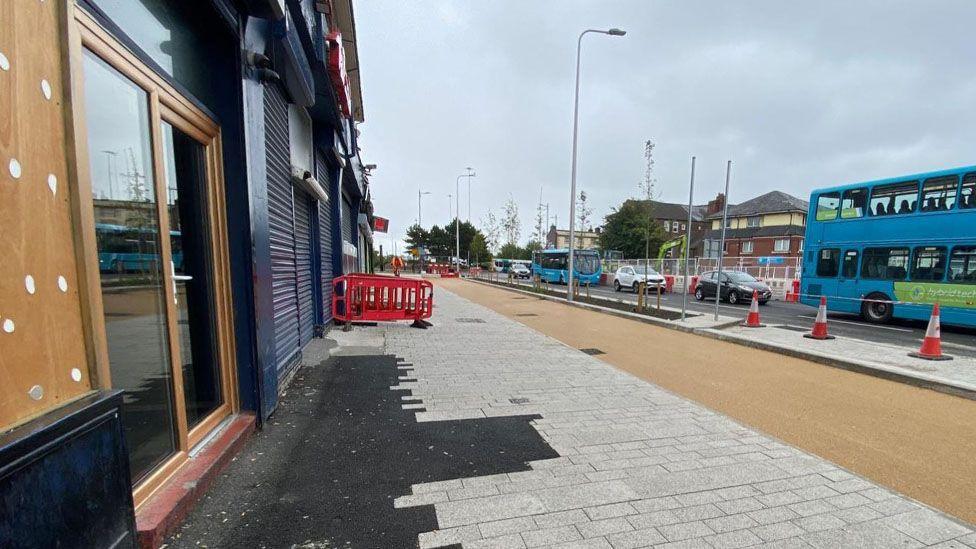 A paved partially paved area outside shops which unfinished roadworks and cones liming the street. Traffic is queuing on the main road.