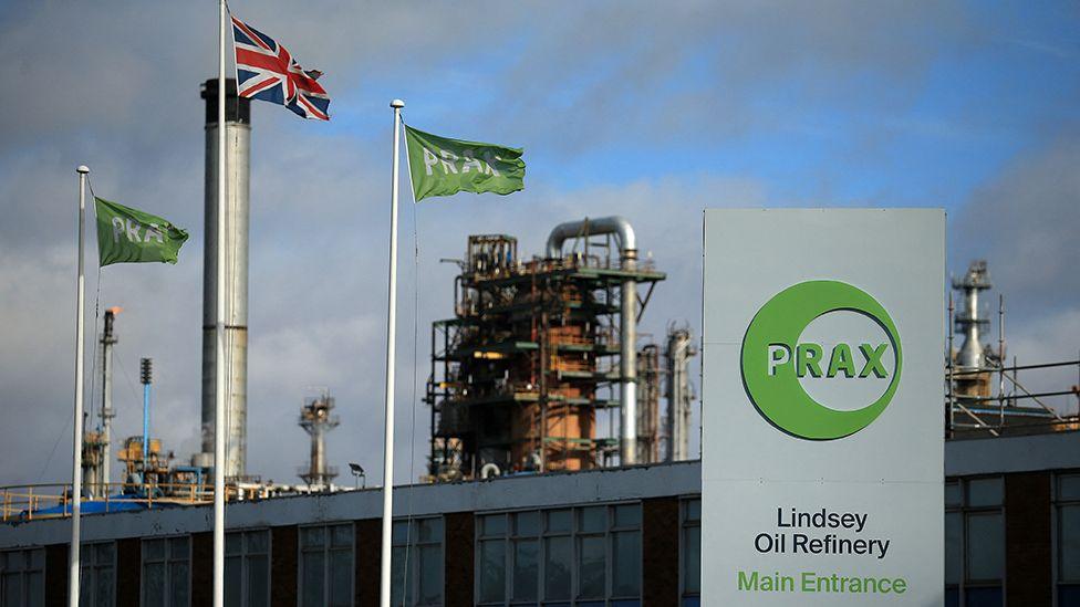 Main entrance of the Prax Lindsey Oil Refinery with a white and green sign and three flag poles with the company's flag and the Union Flag flying in front of the chimneys of the plant