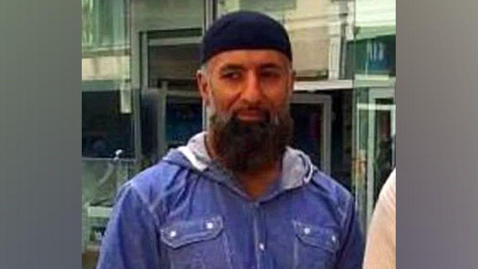 PC Hassan Ali, who has a long dark beard and grey sideburns. He is wearing a blue hooded jacket and is pictured outside.