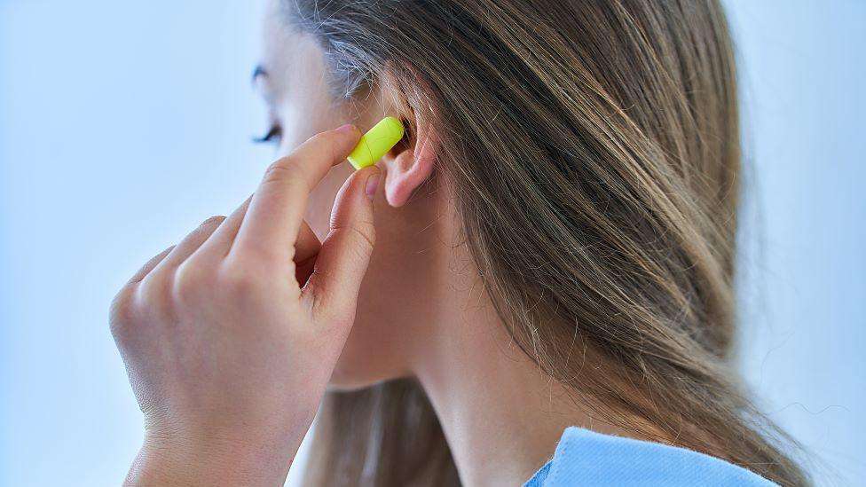 A woman with brunette hair, putting a yellow earplug in her ear.