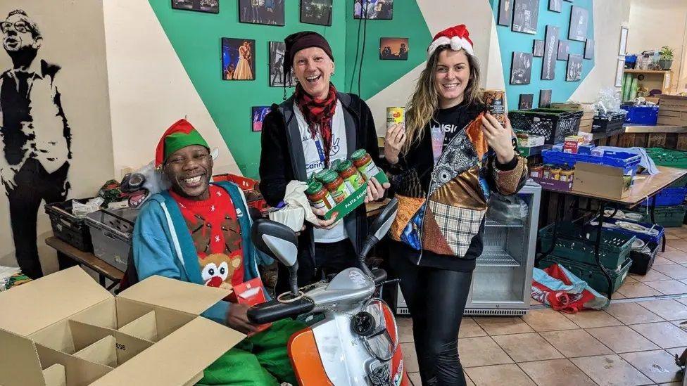 Three people smile at a camera whilst holding food and gifts in their hand. The person on the left is sitting down while the two people on the right are standing up.