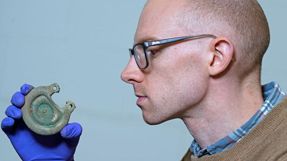 A man with glasses on and a checked shirt closely examines a Bronze Age artefact he is holding in a blue-gloved hand