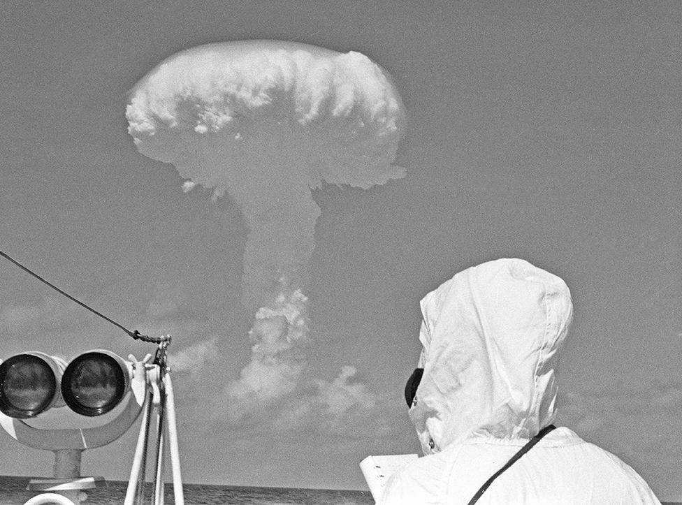 A mushroom cloud rises over the sea while someone looks on from a ship, wearing a white hooded coat and black mask, next to some binoculars. HMS Alert was positioned 35 miles offshore of Malden Island, Kiribati, the site chosen for a series of nuclear tests on 31st May 1957. 