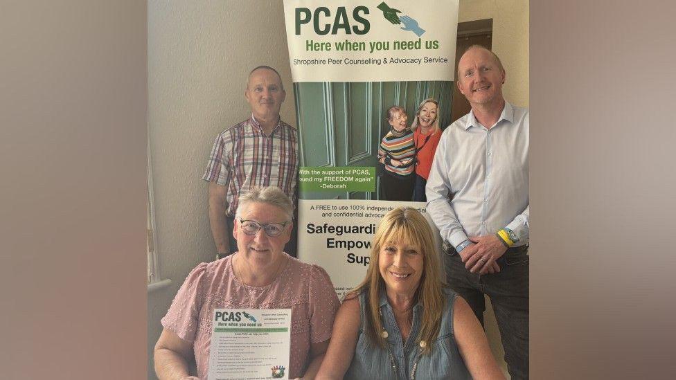 Two women and two men are in front of a large banner that has the PCAS logo on it. It says "here when you need us" and has a picture of two women on it. It also says "safeguarding, empowering, support".
