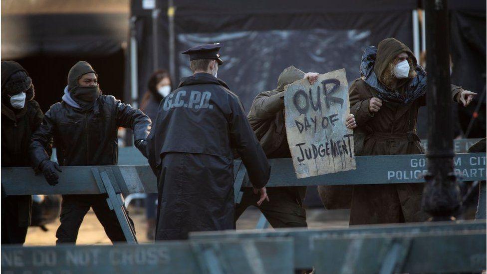 Extras playing anarchists and police officers, the anarchists are behind a police cordon, have their faces covered and are holding signs saying "our day of judgement"