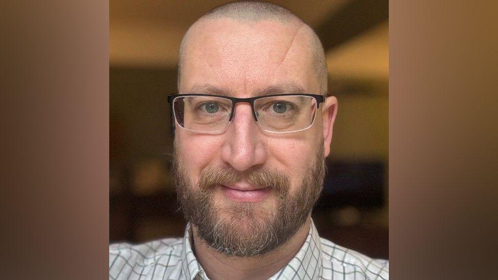 Simon Case in chequered white shirt. He has a beard and square glasses and is looking at the camera. He has a shaved head and is smiling.