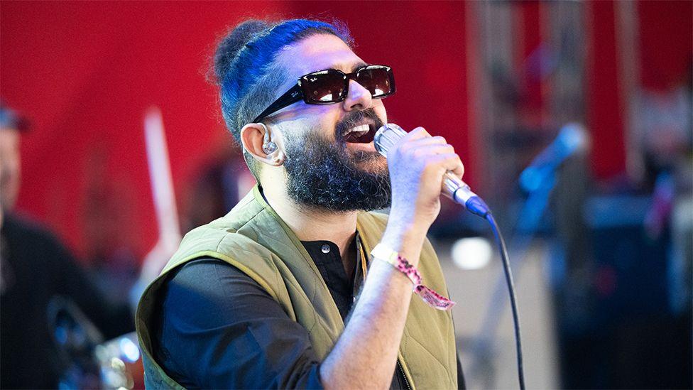 Sid Sriram, a male in black sunglasses, performing on stage at Coachella, with a microphone in his right hand, wearing a green vest jacket with a black top.