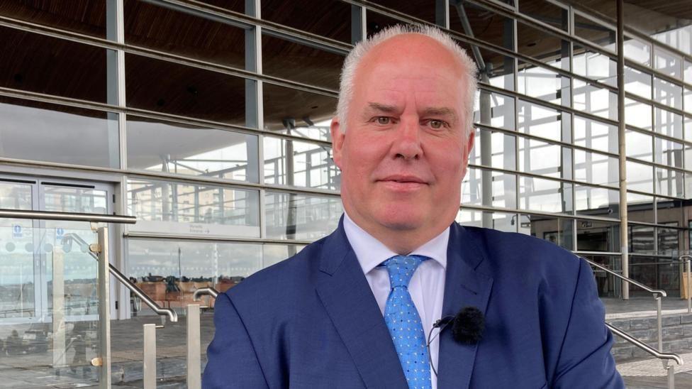 A smiling Andrew RT Davies looking at the camera on the steps in front of the Senedd building in Cardiff Bay, with the many glass panes of the building behind him showing a little of the interior. He is wearing a light blue tie, blue suit, a white shirt and a lapel microphone used for television interviews.