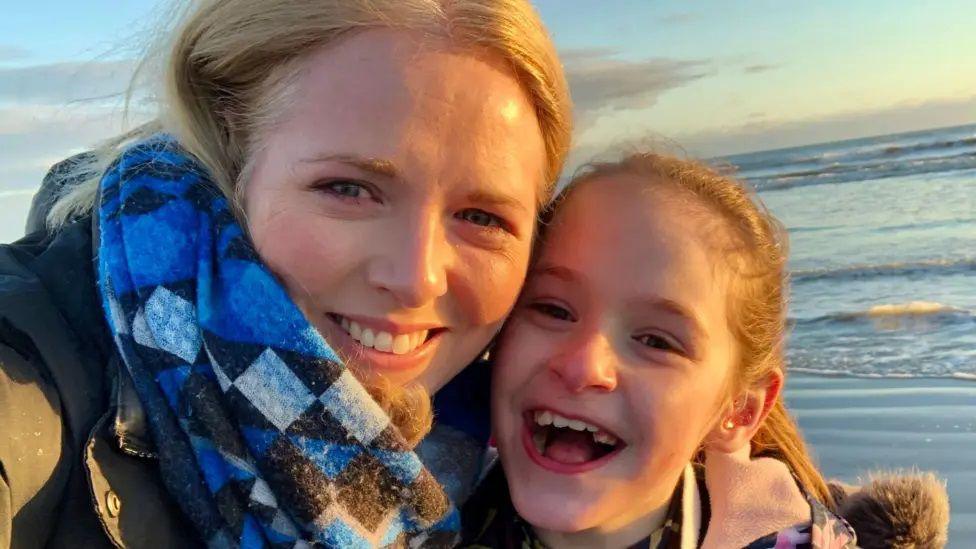 A blonde woman in a blue scarf smiling and looking at the camera while holding her head close to a young girl who is also blonde and smiling with her mouth open.