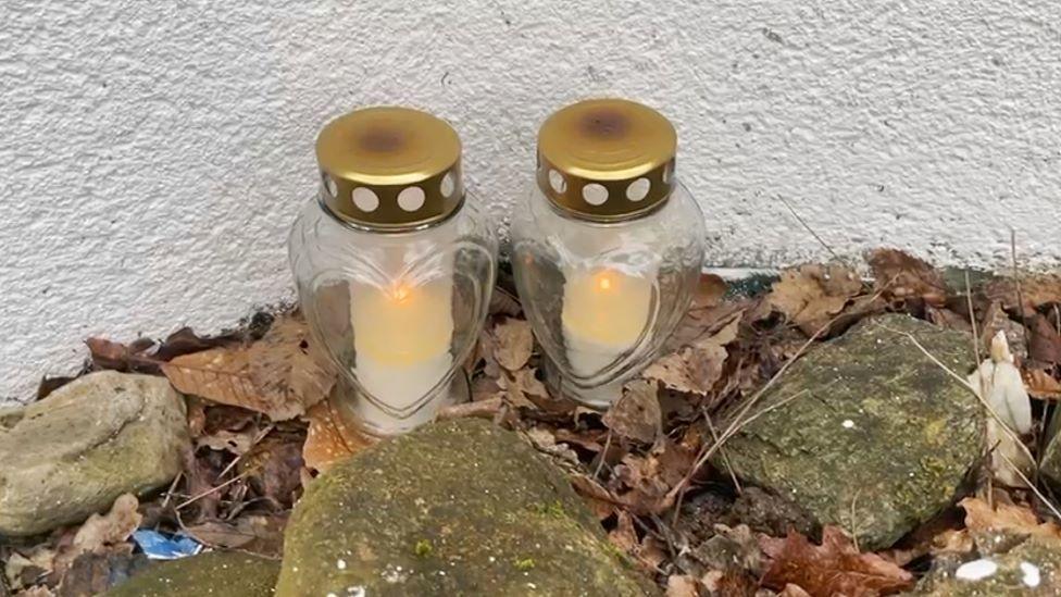 Two candles in heart-shaped glass jars are placed against a white wall, nestled among stones and brown leaves