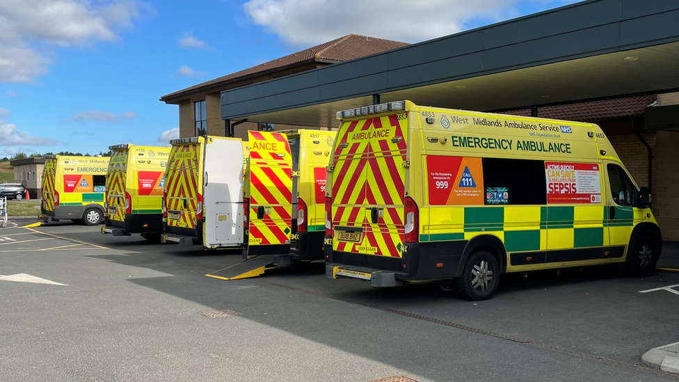 A side-on view of five ambulances parked in a row outside a building. The second one along has its rear doors open and the ramp down.