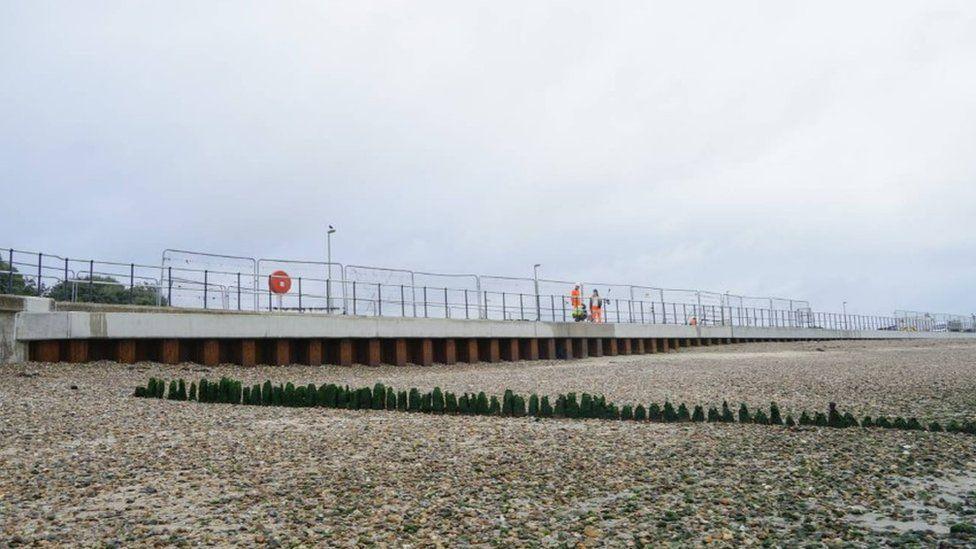 Pebble beach with wooded groyne and metal underneath of seawall up to concrete promenade with railings