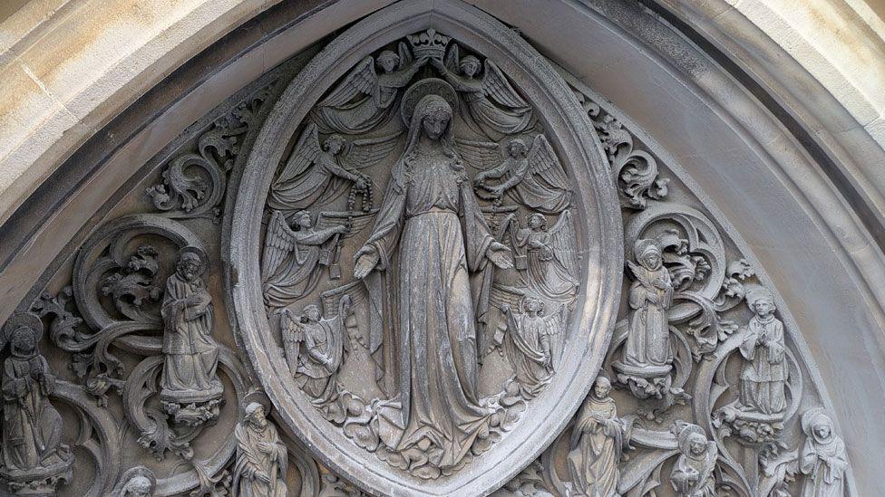 A close up of carving on the outside of the Church of Our Lady and the English Martyrs. In the middle is an oval within which is the Virgin Mary, wearing a long dress, with a halo and surrounded by eight smaller angels. Outside that are carvings of medieval-style foliage with figures of men and women.